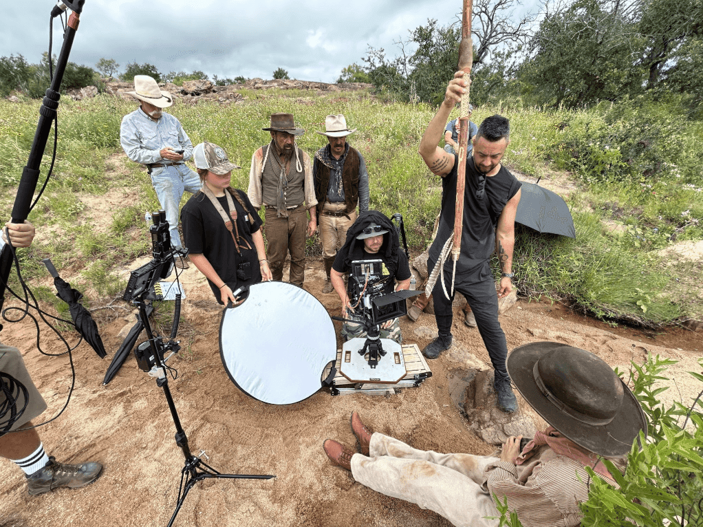 Film crew setting up a creek-side scene with a camera rig and reflector while actors in Western costume wait on a Texas ranch location